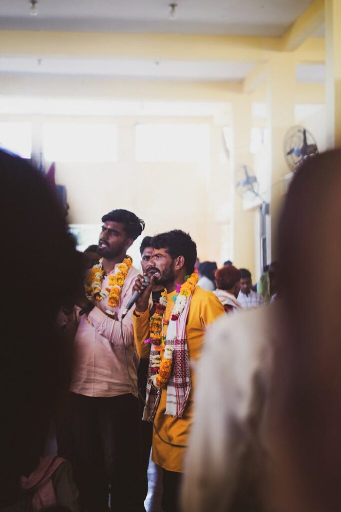 Men adorned with garlands sing joyfully at an indoor Indian celebration, capturing a festive atmosphere.