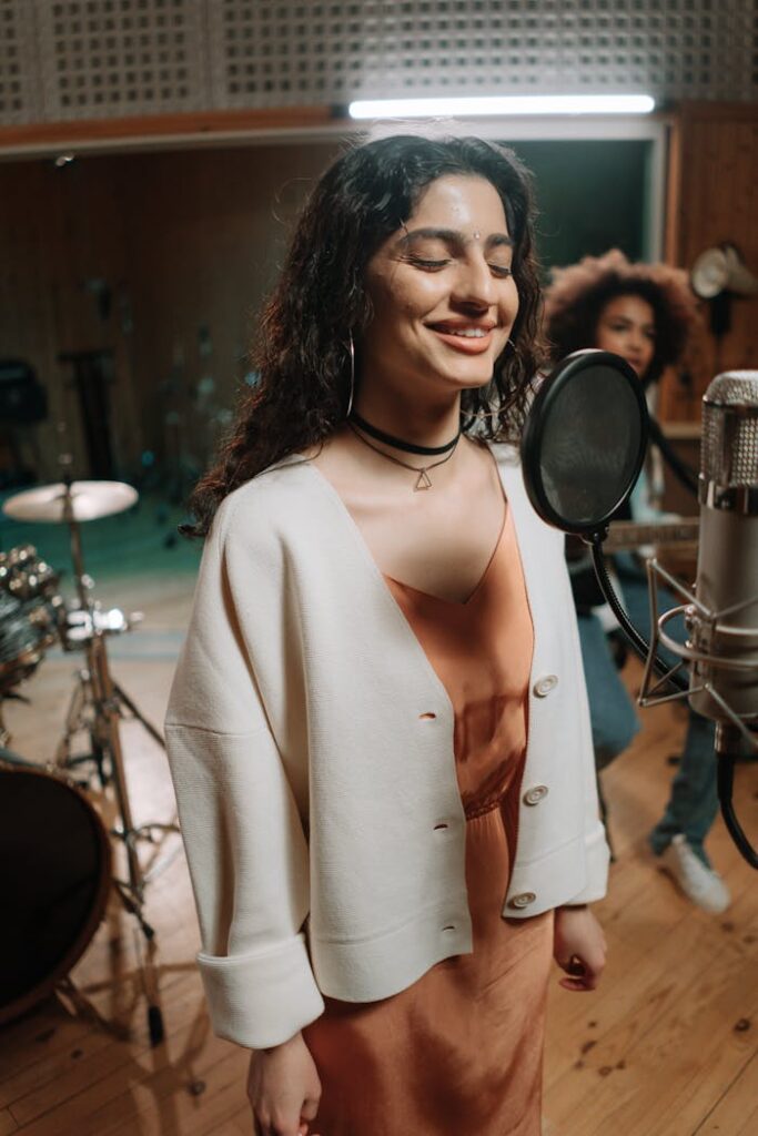 A woman joyfully sings into a condenser microphone in a cozy music studio setting.